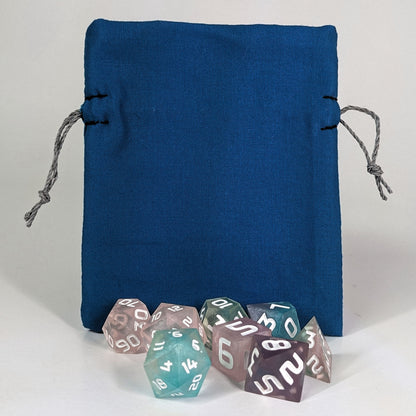 A small, handmade dice bag that is blue stands upright on a white background with a set of colorful dnd dice with white numbers sitting in front of it.