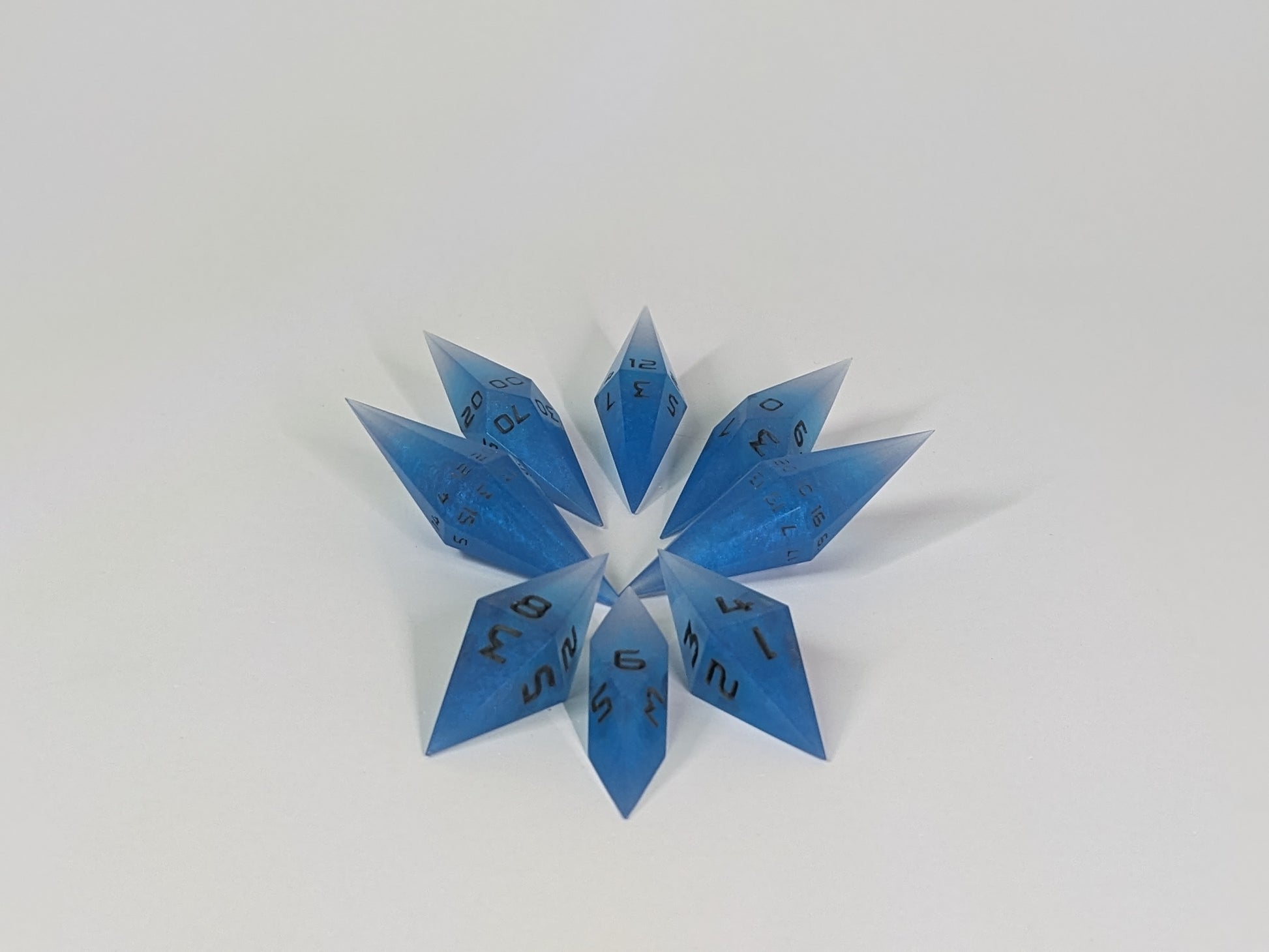 A complete set of resin dice featuring 2 d20s for advantage rolls. These electric blue swirls with grey numbering slender handmade diamond dice are clustered in a tight circle on a white background, highlighting the quality of the artisan dice craftsmanship and the unique pointed shape.