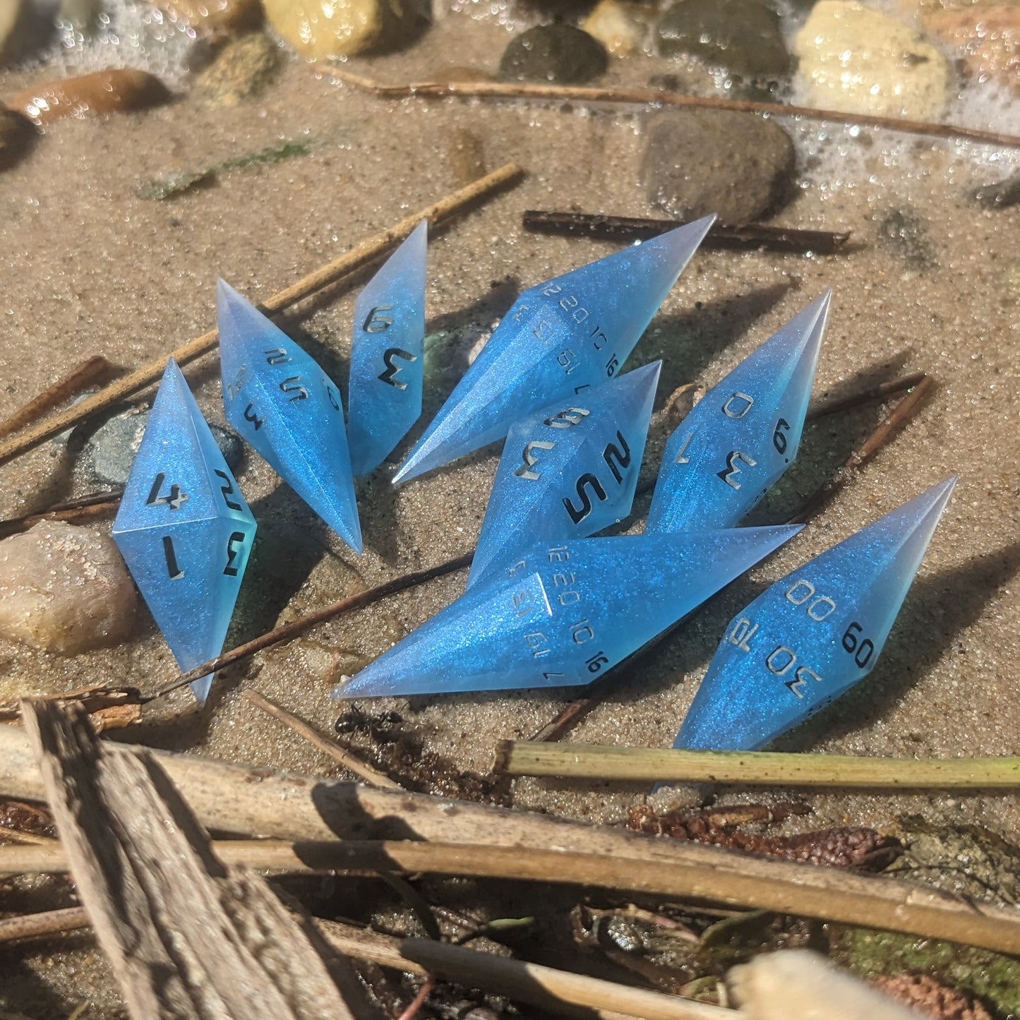 A close-up of electric blue swirled handmade dice with grey numbering resting on a sunny beach. This Lucky Rabbit Dice set of resin dice features a sharp diamond shape, with 2 d20s standing out against the sand, perfectly capturing the vibrant coastal vibe of these artisan dice.