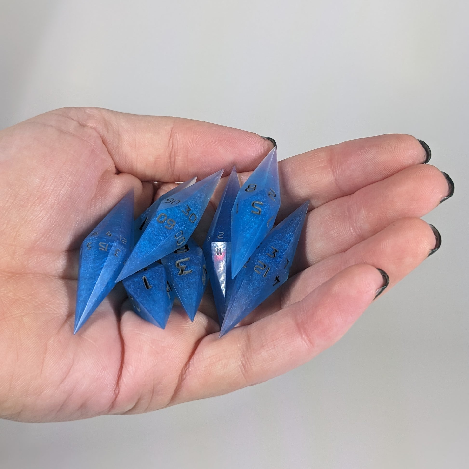 Close-up of electric blue swirls with grey numbering artisan dice resting in a palm on a seamless white backdrop. The handmade dice feature a distinct sharp diamond shape, with 2 d20s visible among the high-quality resin dice.
