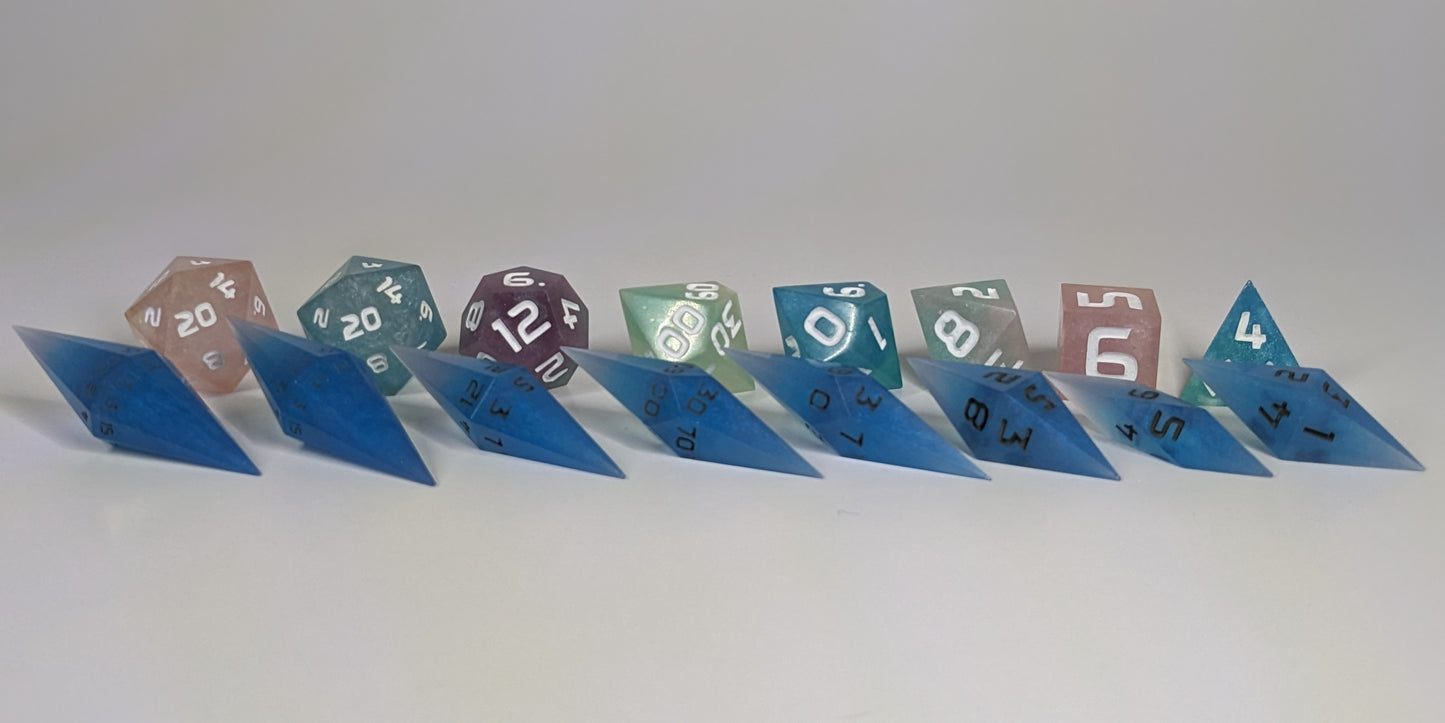 A size comparison of electric blue swirls with grey numbering long pointed diamond handmade dice with standard shapes placed behind for scale. The photo emphasizes the length of the diamond dice being much greater than a standard set as well as the diamond set being much more slender. This artisan dice set features 2 d20s and rests on a seamless white background to highlight the elongated profile of the resin dice.