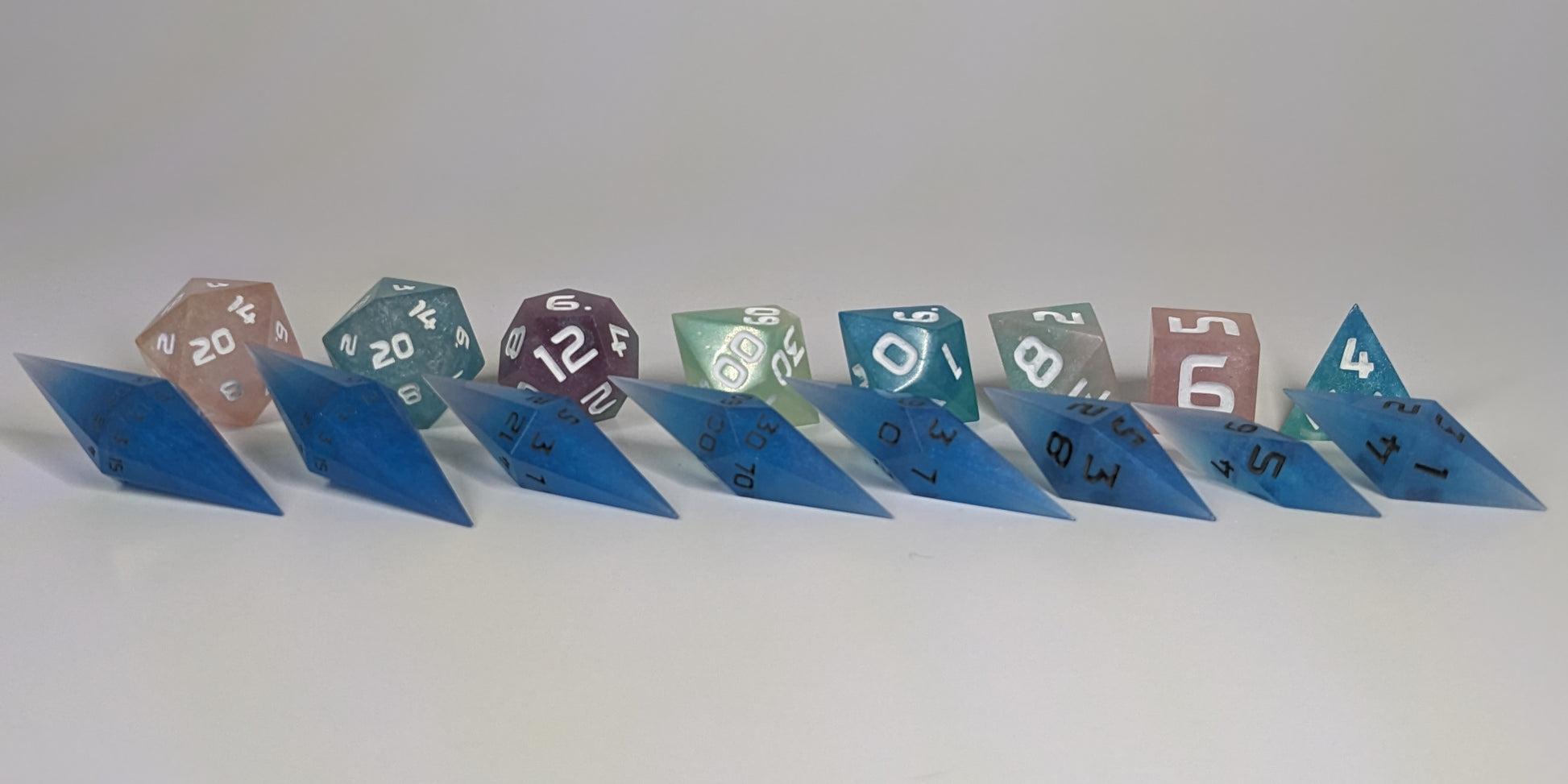 A size comparison of electric blue swirls with grey numbering long pointed diamond handmade dice with standard shapes placed behind for scale. The photo emphasizes the length of the diamond dice being much greater than a standard set as well as the diamond set being much more slender. This artisan dice set features 2 d20s and rests on a seamless white background to highlight the elongated profile of the resin dice.