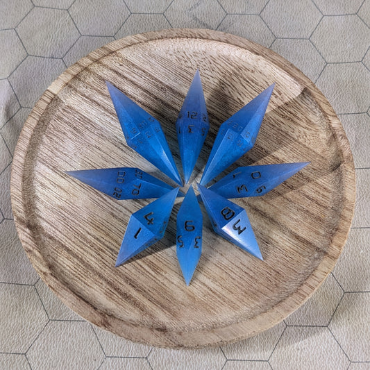 A set of electric blue swirls with grey numbering handmade dice arranged in a wooden circle organizer. The resin dice feature a unique pointed diamond shape with the tips facing outward, highlighting the sharp edges of the two d20 and other artisan dice in the collection. 