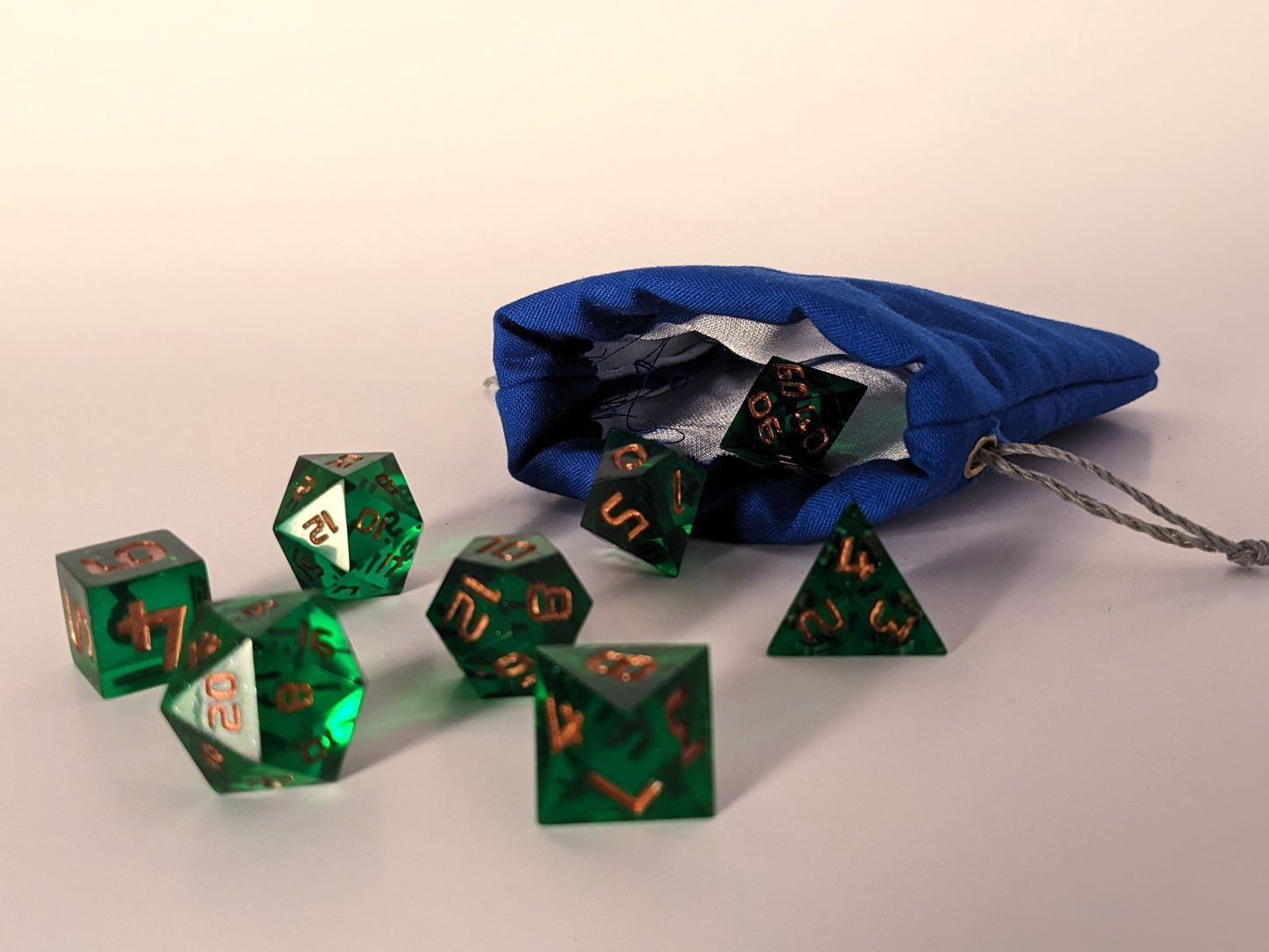 Small handmade dice bag that is blue lays flat with the top open and green dnd dice spilling out from it.