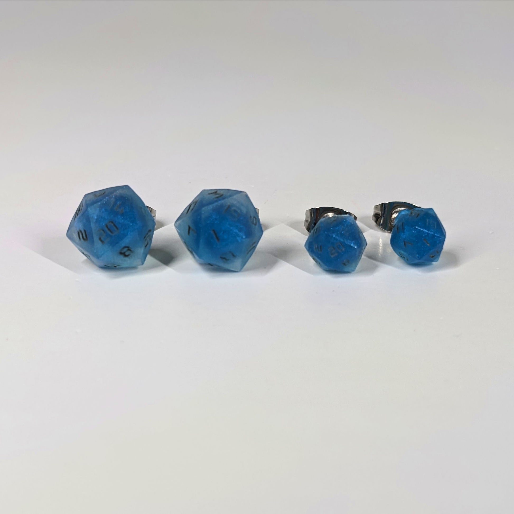 Two pairs of handmade d20 earrings that are swirls of blue with stormy gray numbers on a white background sit in a line with the larger pair on the left and the smaller pair on the right. The earrings on the left of each pair centers the 20 face and the earrings on the right of each pair centers the 1 face.
