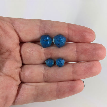 Two pairs of handmade d20 earrings that are swirls of blue with stormy gray numbers held between the fingers of a flat hand. The larger pair is between the pointer and middle finger, the smaller between the middle and ring finger. The earrings on the left of each pair centers the 20 face and the earrings on the right of each pair centers the 1 face.