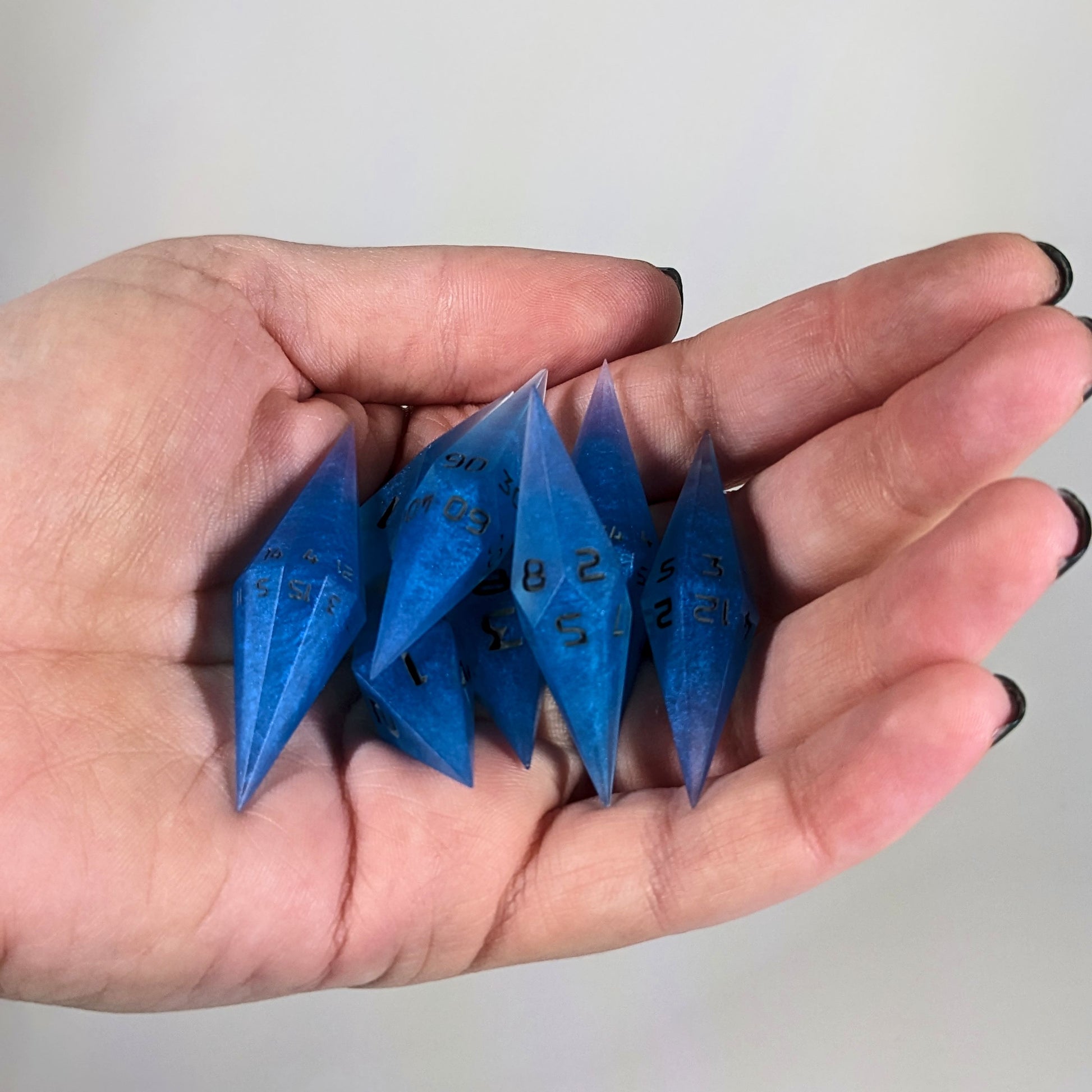An 8 dice set of diamond style dice that are swirls of electric blue with grey numbers are held in a hand. The image shows that the set fits easily in a hand, making them overall smaller, though sharper, than a standard set of dnd dice.