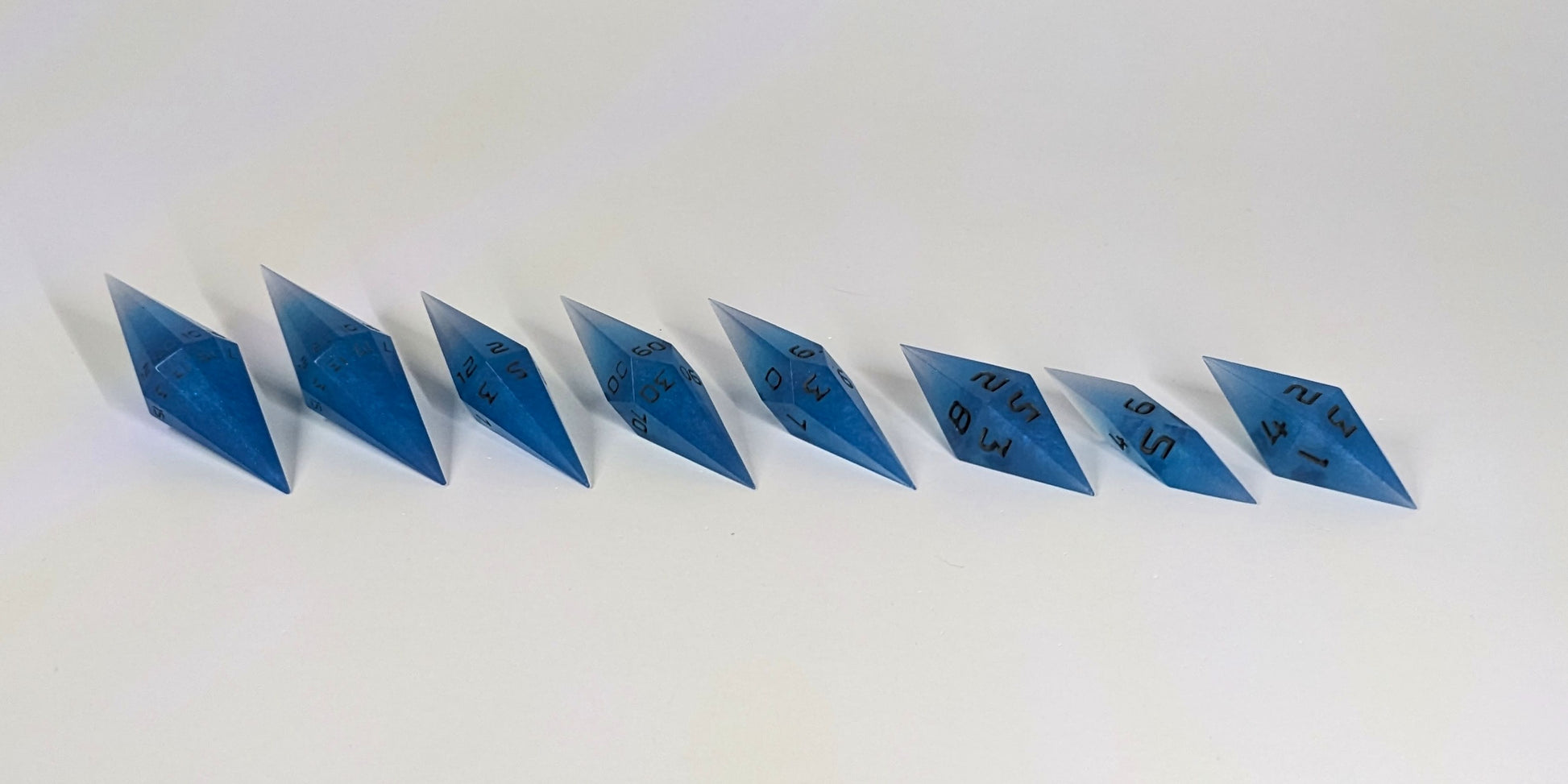A mid angle photo of a complete set of handmade diamond DnD dice that are swirls of electric blue with grey numbers arranged in a straight line with bottom points angled slightly forwards and towards the right. They are lined up with the 2 d20s on the left going down to the d4 on the right side. The highest value number of each die is facing forward.