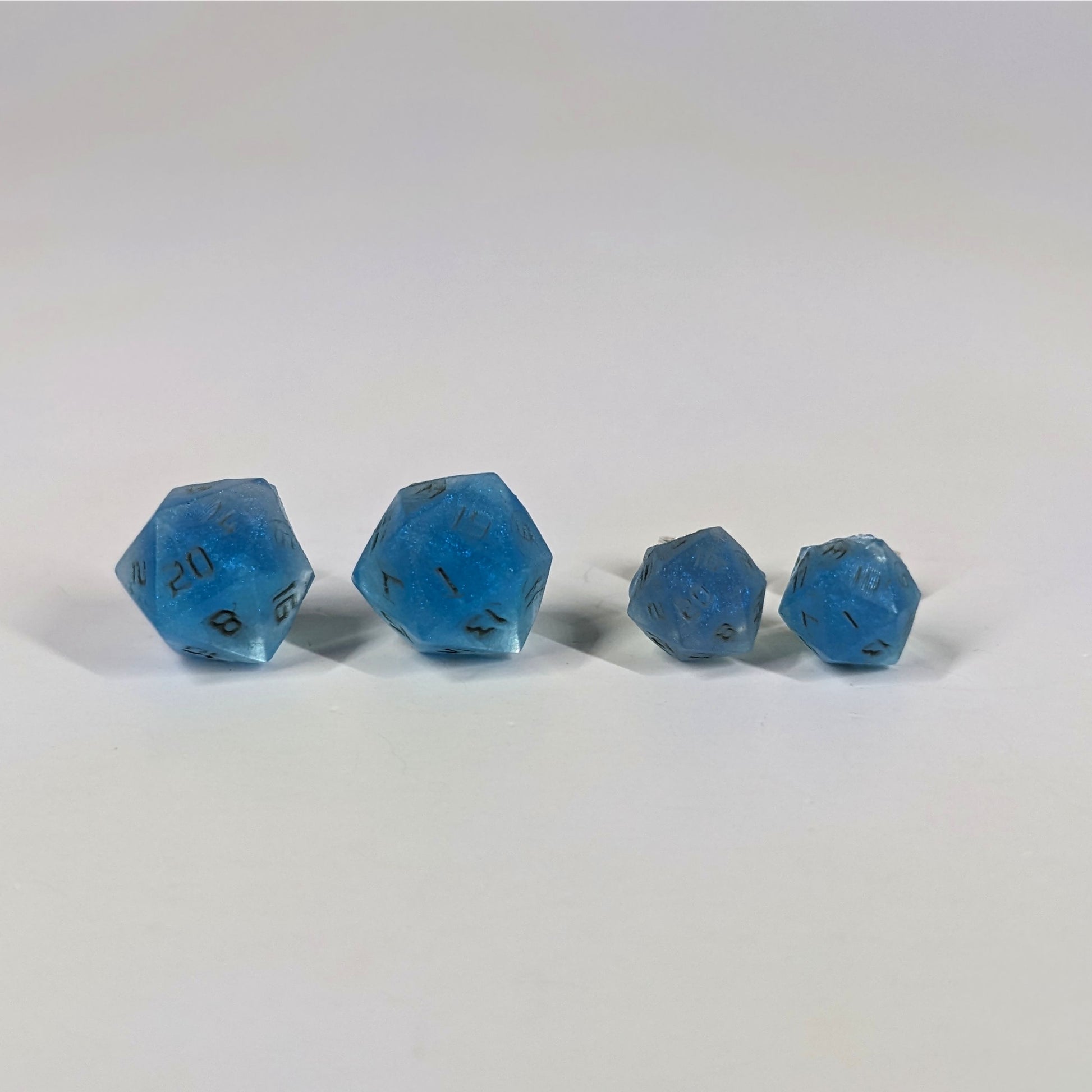 Two pairs of handmade d20 earrings with sterling silver hardware that are swirls of blue with stormy gray numbers on a white background sit in a line with the larger pair on the left and the smaller pair on the right. The earrings on the left of each pair centers the 20 face and the earrings on the right of each pair centers the 1 face.