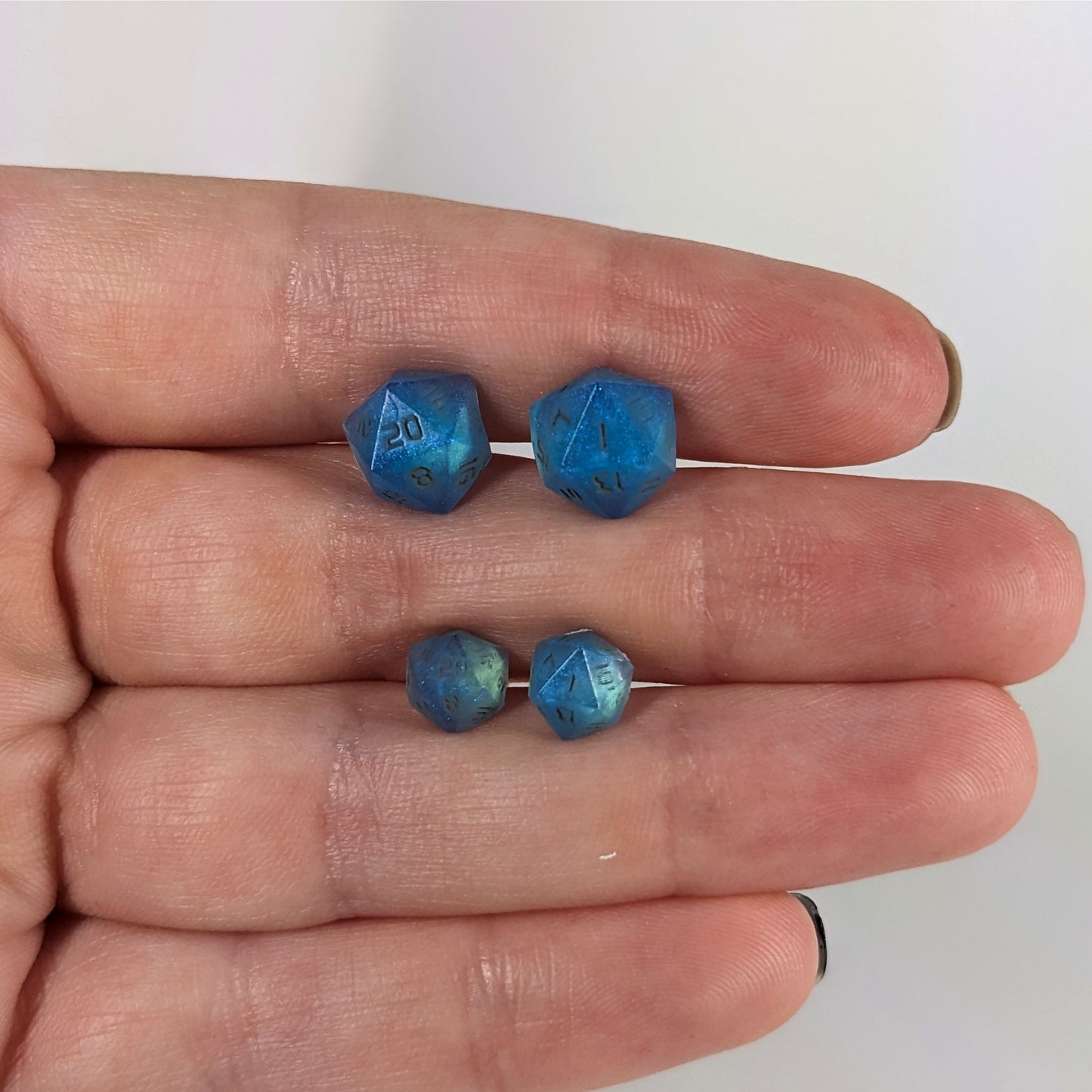 Two pairs of handmade d20 earrings with sterling silver hardware that are swirls of blue with stormy gray numbers held between the fingers of a flat hand. The larger pair is between the pointer and middle finger, the smaller between the middle and ring finger. The earrings on the left of each pair centers the 20 face and the earrings on the right of each pair centers the 1 face.