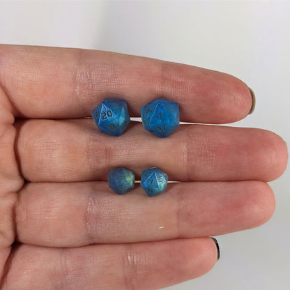 Two pairs of handmade d20 earrings with sterling silver hardware that are swirls of blue with stormy gray numbers held between the fingers of a flat hand. The larger pair is between the pointer and middle finger, the smaller between the middle and ring finger. The earrings on the left of each pair centers the 20 face and the earrings on the right of each pair centers the 1 face.