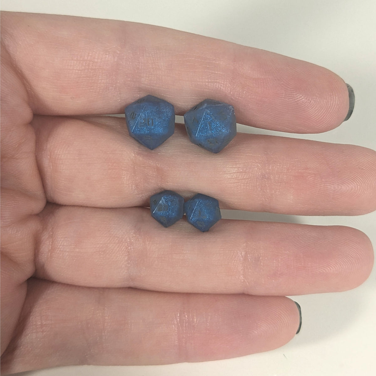 Two pairs of handmade d20 earrings with titanium hardware that are swirls of blue with stormy gray numbers held between the fingers of a flat hand. The larger pair is between the pointer and middle finger, the smaller between the middle and ring finger. The earrings on the left of each pair centers the 20 face and the earrings on the right of each pair centers the 1 face.