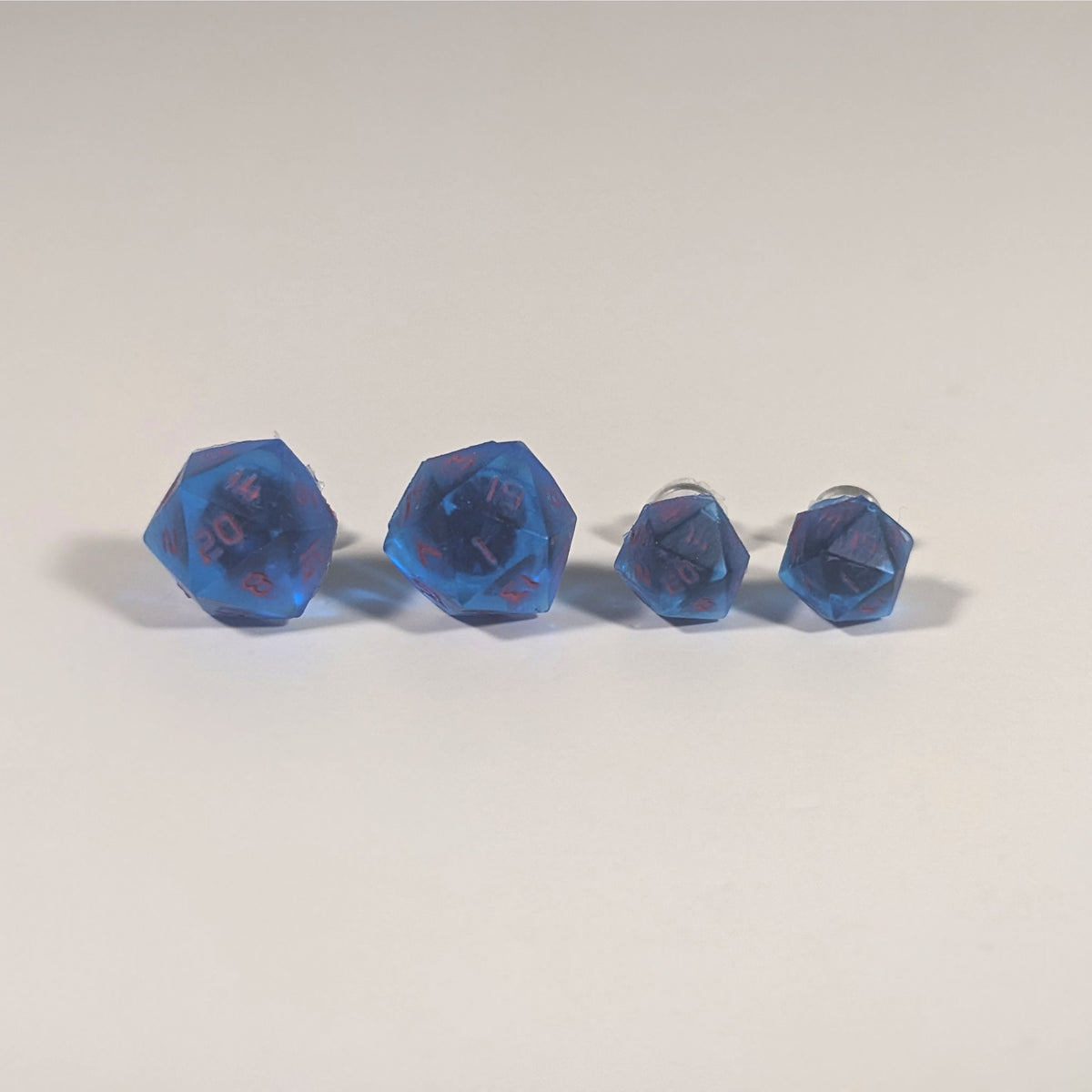 Two pairs of handmade d20 earrings with titanium hardware that are rich blue with bright pink numbers on a white background sit in a line with the larger pair on the left and the smaller pair on the right. The earrings on the left of each pair centers the 20 face and the earrings on the right of each pair centers the 1 face.
