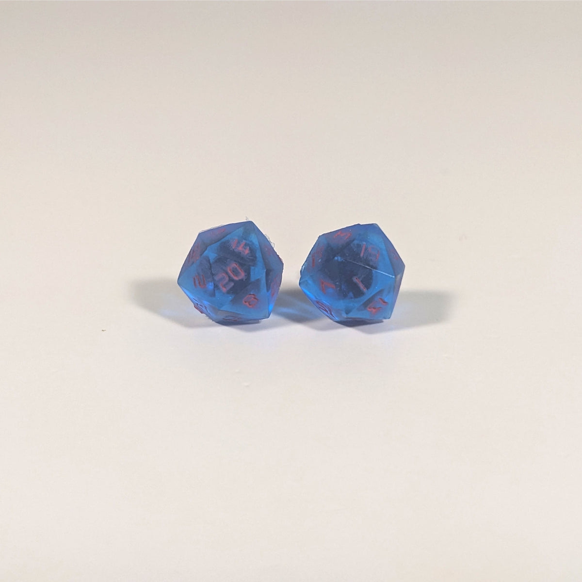 A single pair of the larger size handmade d20 earrings with titanium hardware that are rich blue with bright pink numbers on a white background. The earring on the left centers the 20 face and the earring on the right centers the 1 face.