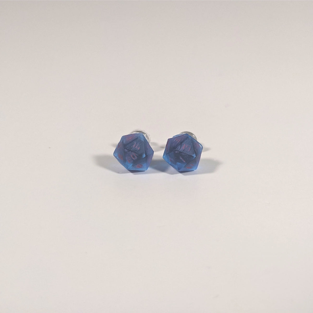 A single pair of the smaller size handmade d20 earrings with titanium hardware that are rich blue with bright pink numbers on a white background. The earring on the left centers the 20 face and the earring on the right centers the 1 face.