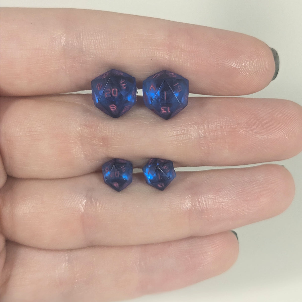 Two pairs of handmade d20 earrings with titanium hardware that are rich blue with bright pink numbers held between the fingers of a flat hand. The larger pair is between the pointer and middle finger, the smaller between the middle and ring finger. The earrings on the left of each pair centers the 20 face and the earrings on the right of each pair centers the 1 face.