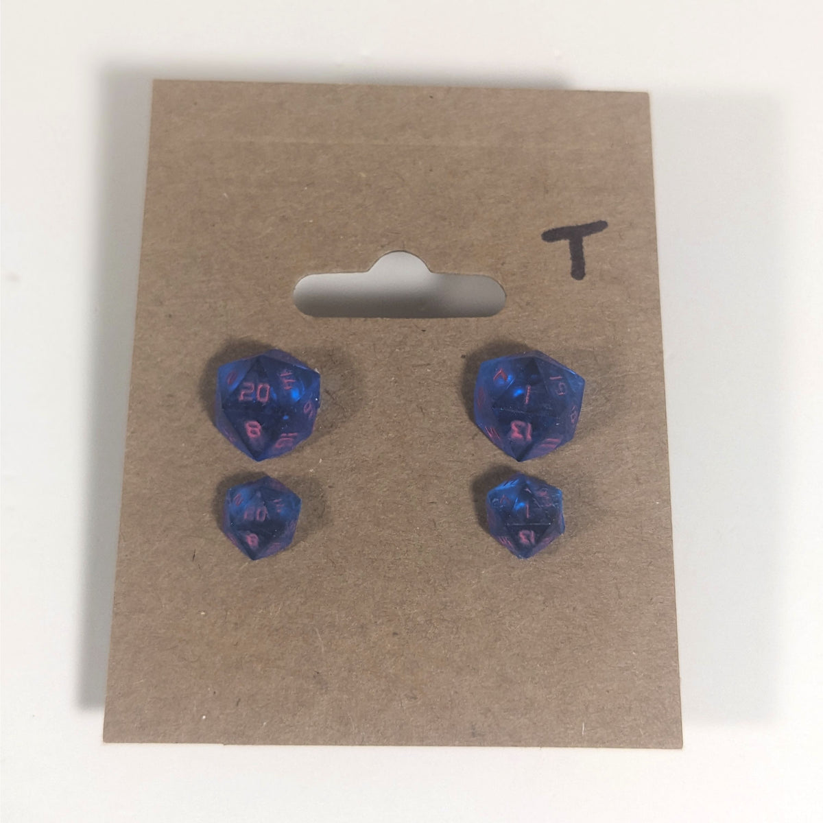 Two pairs of handmade d20 earrings with titanium hardware that are rich blue with bright pink numbers on a kraft brown earring card with a T in the top right corner. The earrings on the left of each pair centers the 20 face and the earrings on the right of each pair centers the 1 face.