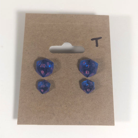 Two pairs of handmade d20 earrings with titanium hardware that are rich blue with bright pink numbers on a kraft brown earring card with a T in the top right corner. The earrings on the left of each pair centers the 20 face and the earrings on the right of each pair centers the 1 face.