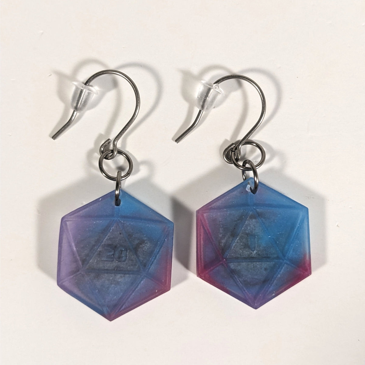 A pair of blue, purple, and silver dangly, flat d20 earrings with silver color titanium and niobium hardware and bell shaped rubber backs on a white background. The earring on the left has a 20 on it and the earring on the right has a 1 on it, both in a sci-fi style font.
