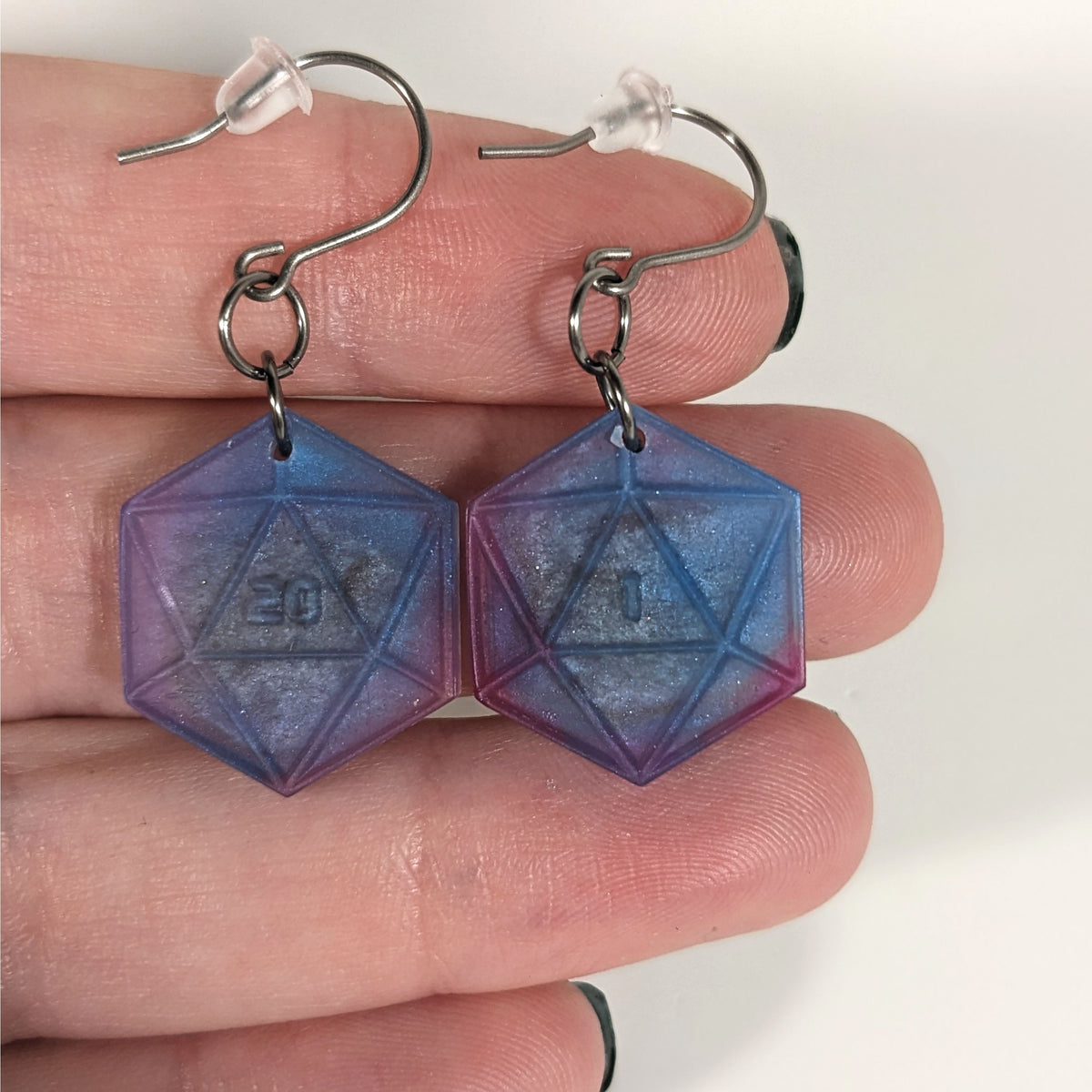 A pair of blue, purple, and silver dangly, flat d20 earrings with silver color titanium and niobium hardware with bell shaped rubber backs held on on a hand on a white background. The earring on the left has a 20 on it and the earring on the right has a 1 on it, both in a sci-fi style font.