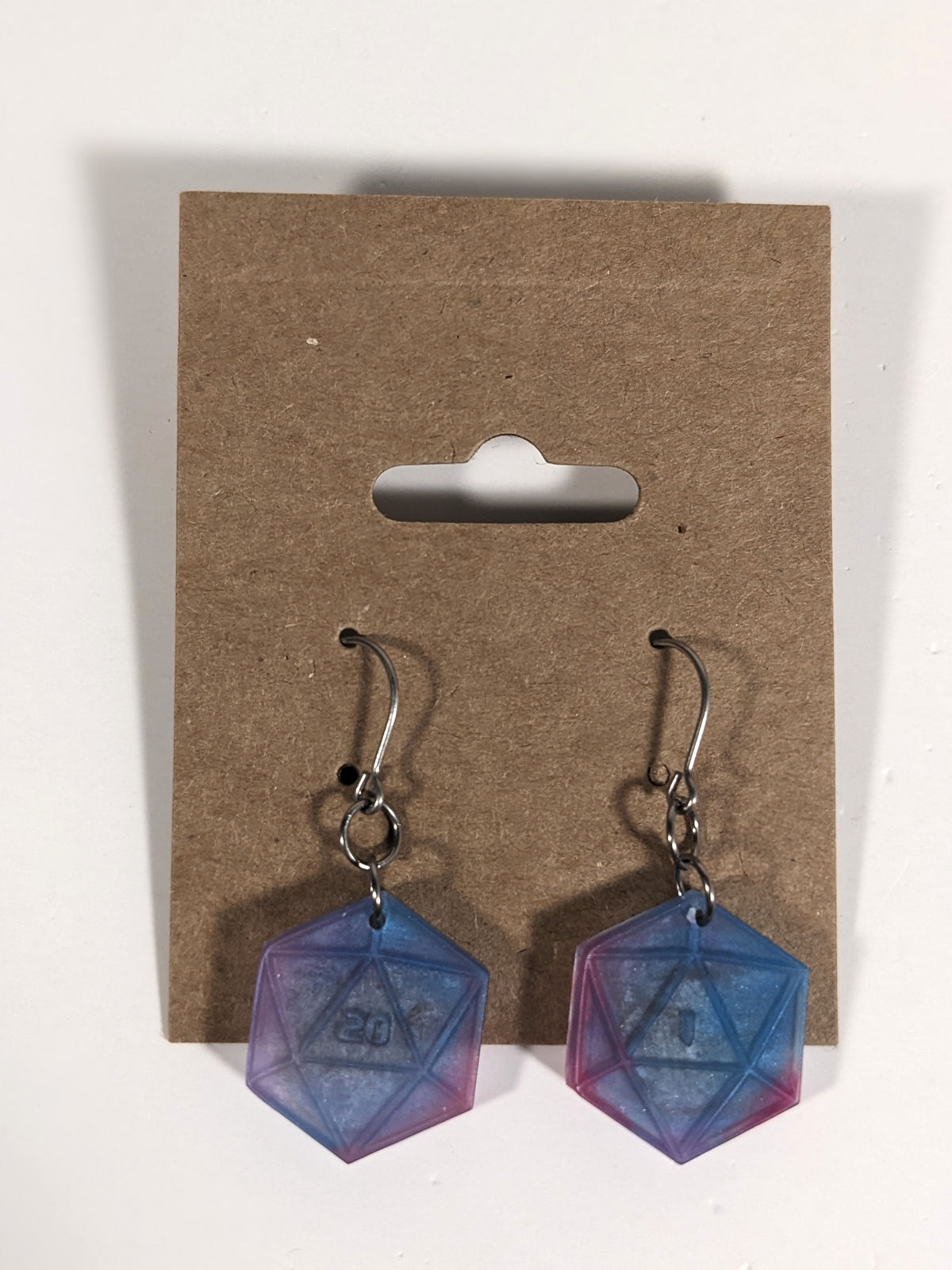 A pair of purple, blue, and silver dangly, flat d20 earrings with silver color titanium and niobium hardware on a kraft earring card on a white background. The earring on the left has a 20 on it and the earring on the right has a 1 on it, both in a sci-fi style font.