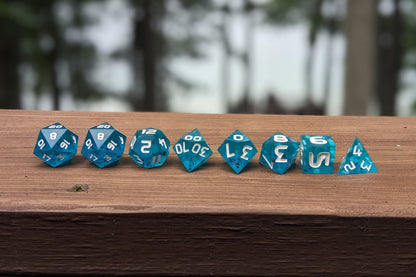 A straight on photo of a complete set of polyhedral dice that are  blue with red flakies and white numbers arranged in a straight line on a wooden railing. They are lined up with the 2 d20s on the left going down to the d4 on the right side. The highest value number of each die is facing up.