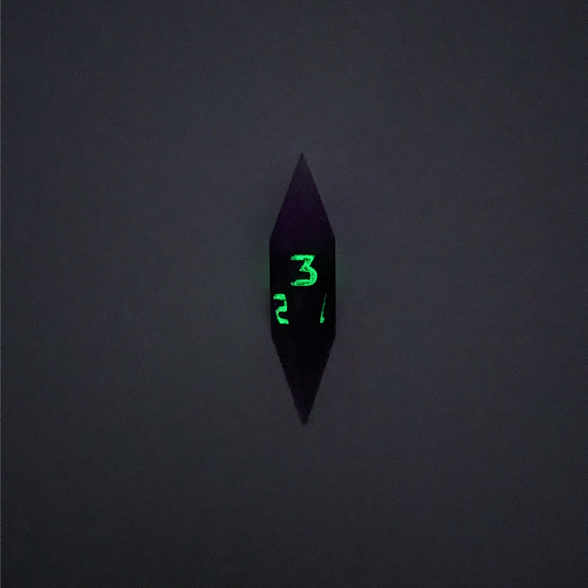 A high angle view of a diamond d3 with swirls of purple and green with black numbers that glow in the dark on a white background. This photo is taken in the dark to show the black numbers glowing green in the dark.