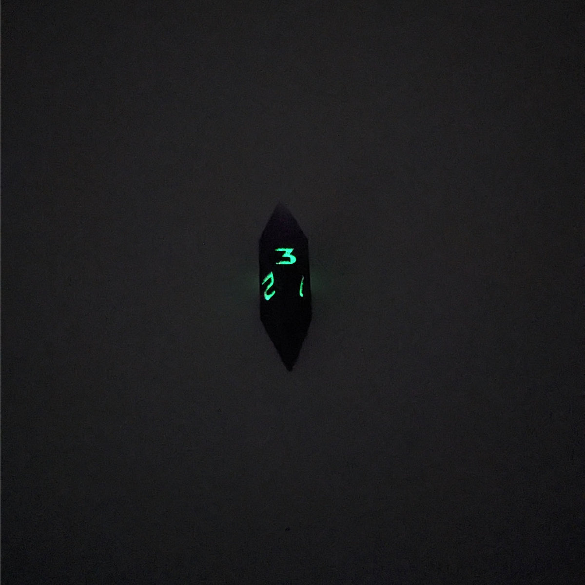 A mid angle view of a diamond d3 with swirls of purple and green with black numbers that glow in the dark on a white background. This photo was taken in the dark to show the black numbers glowing green in the dark.