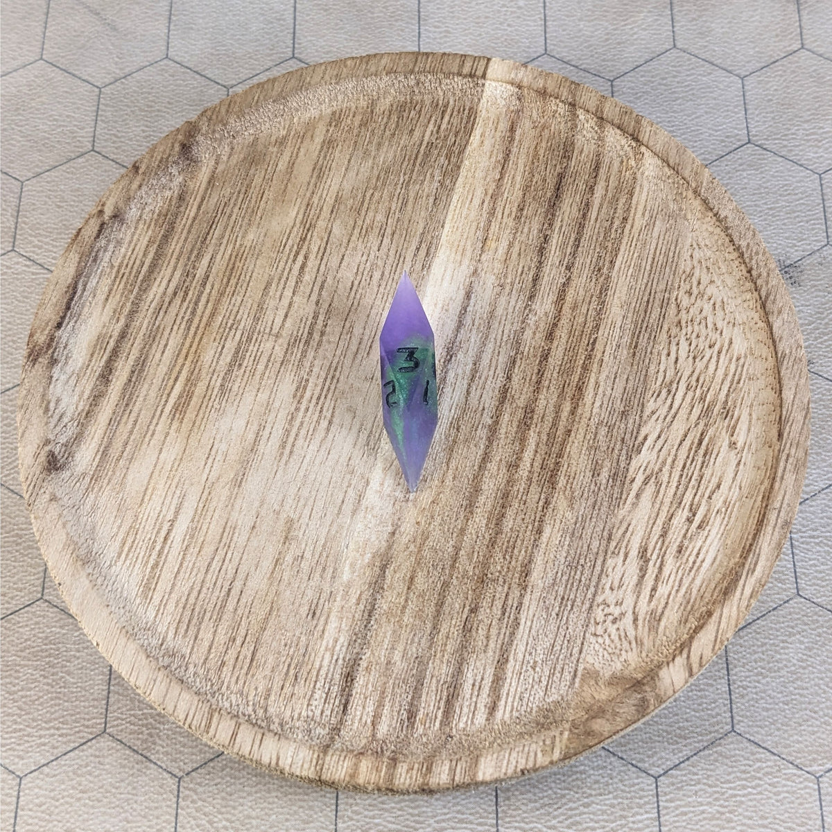 A high angle view of a diamond d3 with swirls of purple and green with black numbers that glow in the dark. It is sitting in the center of a wooden circle.