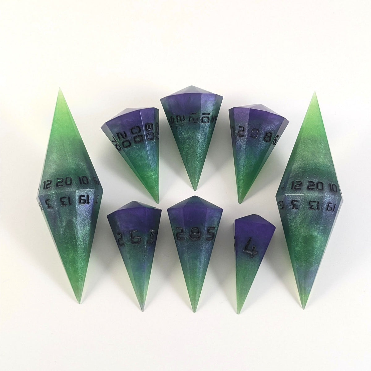 A dagger style DnD dice set that is  green and purple with black numbers that glow in the dark is arranged with a d20 each on the right and left, with the d%, d12, and d10 along the top and the d6, d8, and d4 along the bottom, all in line with the d20s.