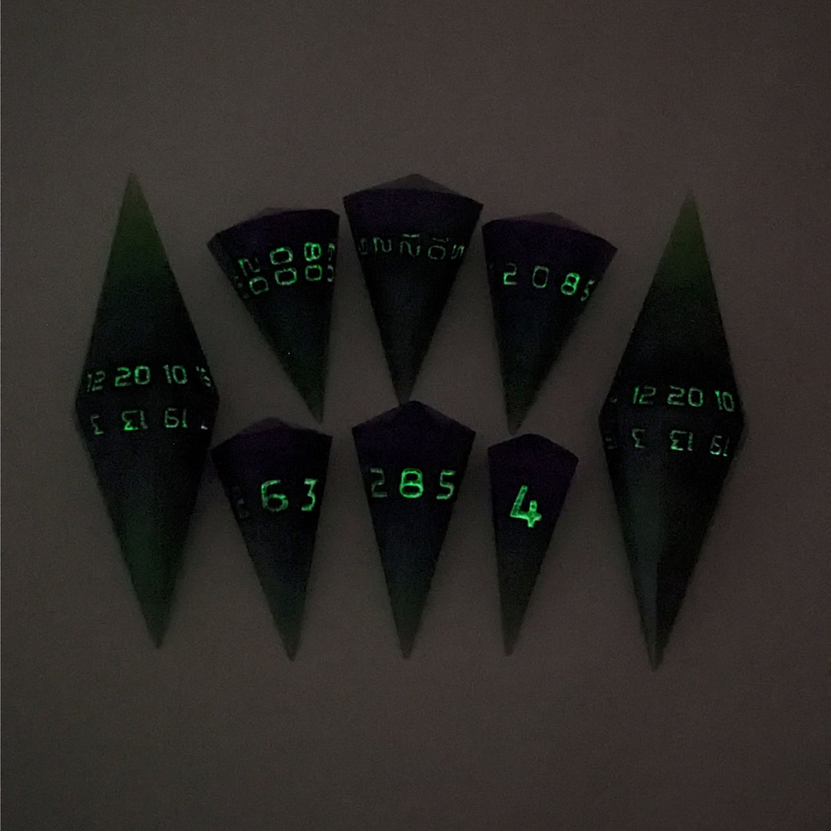 A dagger style DnD dice set that is  green and purple with black numbers that glow in the dark is arranged with a d20 each on the right and left, with the d%, d12, and d10 along the top and the d6, d8, and d4 along the bottom, all in line with the d20s. The photo was taken in the dark to show the black numbers glowing green.