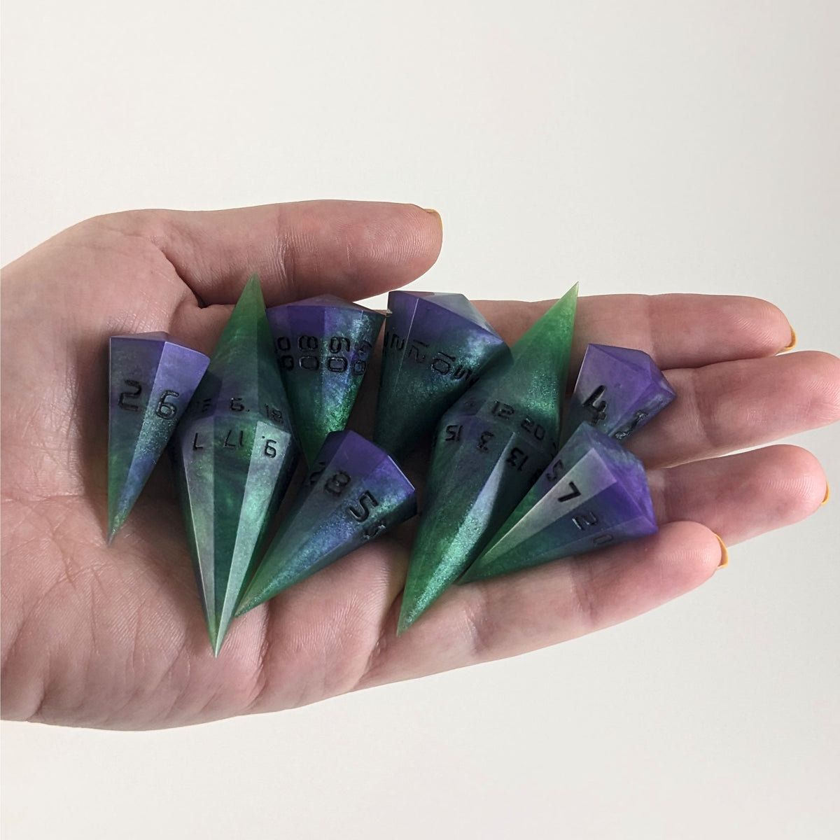An 8 dice set of dagger style dice that are green and purple with black numbers that glow in the dark are held in a hand. The image shows that the set just fits in a hand, making them overall larger than a standard set of dnd dice.