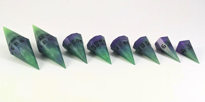A straight on photo of a complete set of dagger DnD dice that are green and purple with black numbers that glow in the dark arranged in a straight line with points angled toward the right. They are lined up with the 2 d20s on the left going down to the d4 on the right side. The highest value number of each die is facing forward.