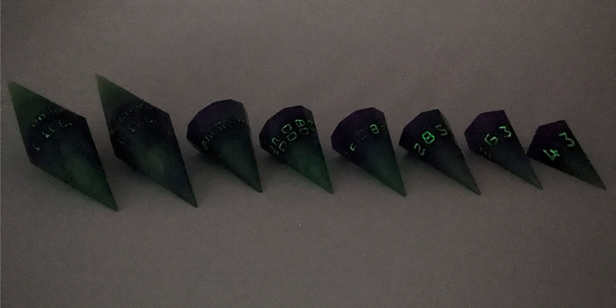 A straight on photo of a complete set of dagger DnD dice that are purple and green with black numbers that glow in the dark arranged in a straight line with points angled toward the right. They are lined up with the 2 d20s on the left going down to the d4 on the right side. The highest value number of each die is facing forward. The photo is taken in the dark to show the black numbers glowing green.
