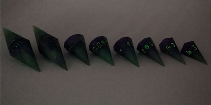 A straight on photo of a complete set of dagger DnD dice that are purple and green with black numbers that glow in the dark arranged in a straight line with points angled toward the right. They are lined up with the 2 d20s on the left going down to the d4 on the right side. The highest value number of each die is facing forward. The photo is taken in the dark to show the black numbers glowing green.