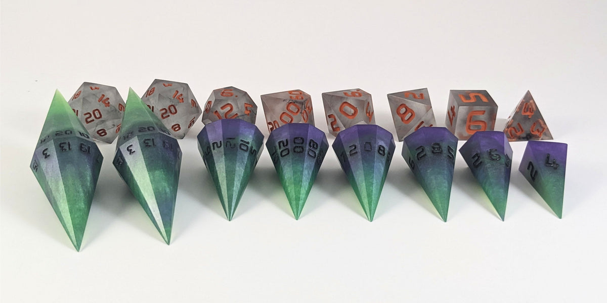 A straight on photo of a complete set of dagger dice that are green and purple with black numbers that glow in the dark arranged in a straight line with points angled forward. They are lined up with the 2 d20s on the left going down to the d4 on the right side. Behind the dagger set is a standard set of polyhedral dnd dice to allow for size comparison. The dagger dice are much larger overall, but not taller. The highest value number of each dagger die is facing up.