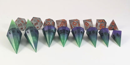 A straight on photo of a complete set of dagger dice that are green and purple with black numbers that glow in the dark arranged in a straight line with points angled forward. They are lined up with the 2 d20s on the left going down to the d4 on the right side. Behind the dagger set is a standard set of polyhedral dnd dice to allow for size comparison. The dagger dice are much larger overall, but not taller. The highest value number of each dagger die is facing up.