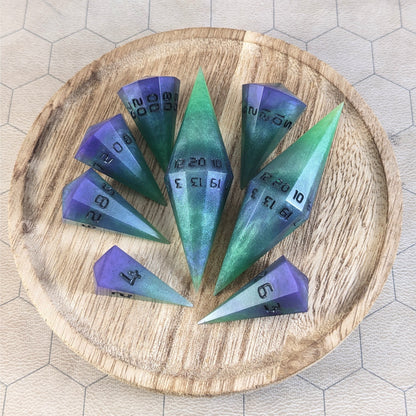 A complete set of dagger style resin dice that are green and purple with black numbers that glow in the dark are arranged on a wooden circle. The 2 d20s are next to each other with the d12 placed between them on top. The d6 is to the bottom right of the d20s and the rest of the dice are arranged along the left side, points angled toward the d20s.