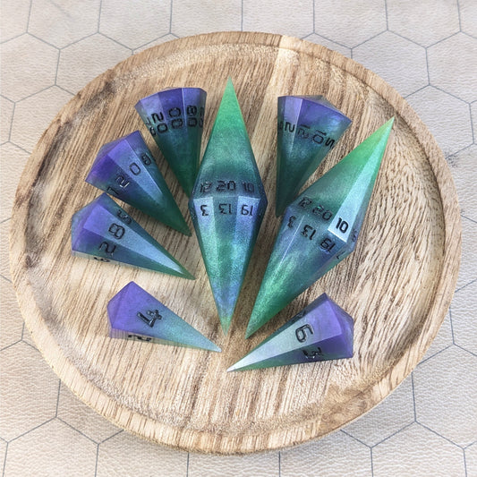 A complete set of dagger style resin dice that are green and purple with black numbers that glow in the dark are arranged on a wooden circle. The 2 d20s are next to each other with the d12 placed between them on top. The d6 is to the bottom right of the d20s and the rest of the dice are arranged along the left side, points angled toward the d20s.