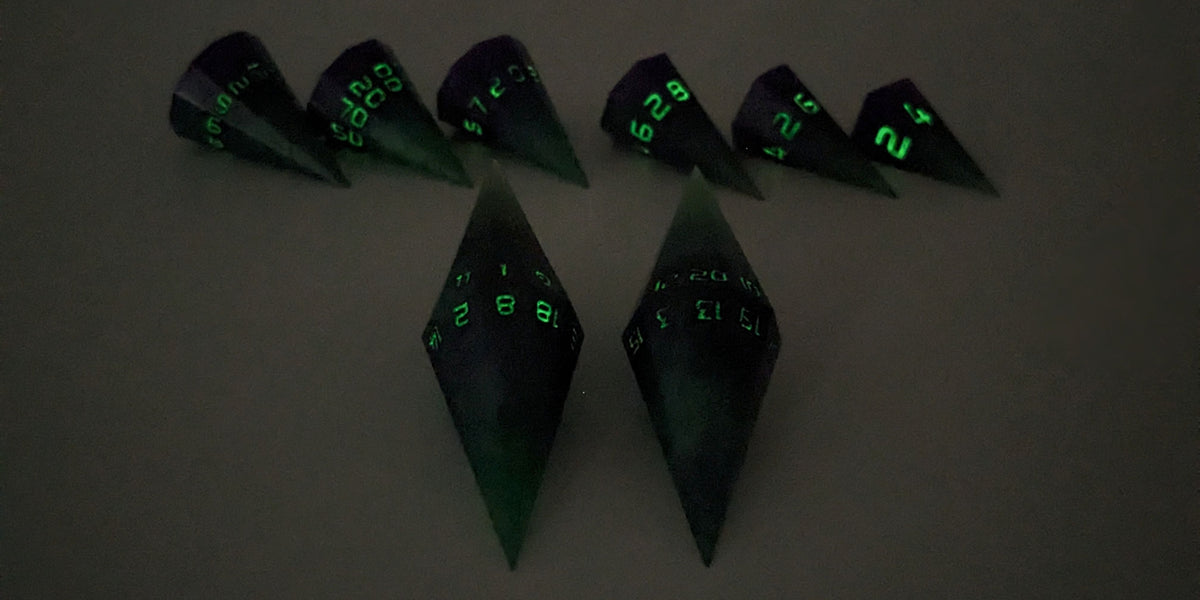 A straight on photo of a dagger set of DnD dice that are green and purple with black numbers that glow in the dark focusing on 2 d20s in the center. The d20 on the left has the 1 face up and the d20 on the right has the 20 face up. The other 6 dice are lined up in the background but are out of focus, while the d20s are in sharp focus. The photo is taken in the dark to show the black numbers glowing green.