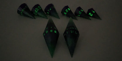 A straight on photo of a dagger set of DnD dice that are green and purple with black numbers that glow in the dark focusing on 2 d20s in the center. The d20 on the left has the 1 face up and the d20 on the right has the 20 face up. The other 6 dice are lined up in the background but are out of focus, while the d20s are in sharp focus. The photo is taken in the dark to show the black numbers glowing green.