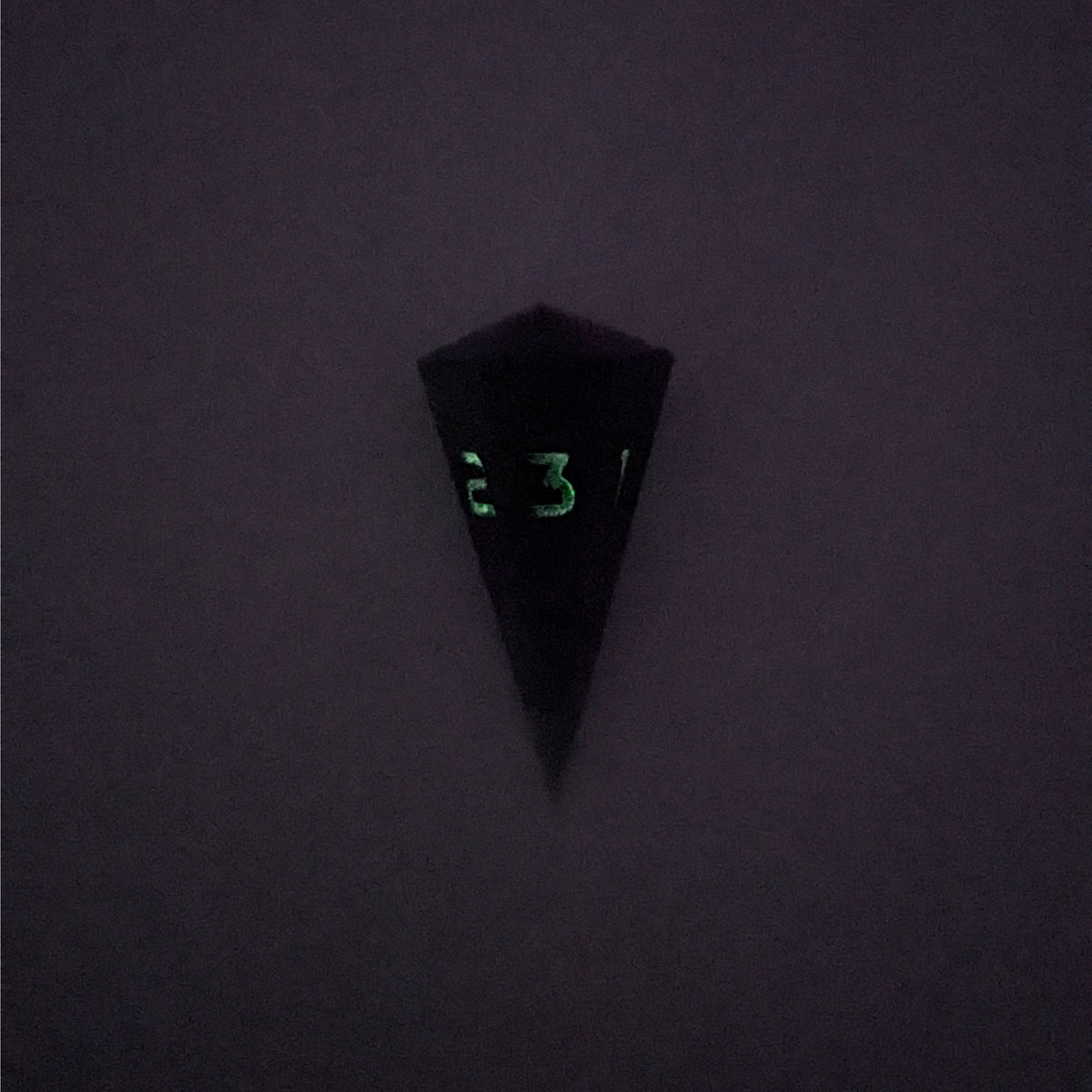 A high angle view of a dagger d3 with swirls of purple and green with black numbers that glow in the dark on a white background. This photo is taken in the dark to show the black numbers glowing green in the dark.