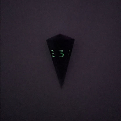 A high angle view of a dagger d3 with swirls of purple and green with black numbers that glow in the dark on a white background. This photo is taken in the dark to show the black numbers glowing green in the dark.