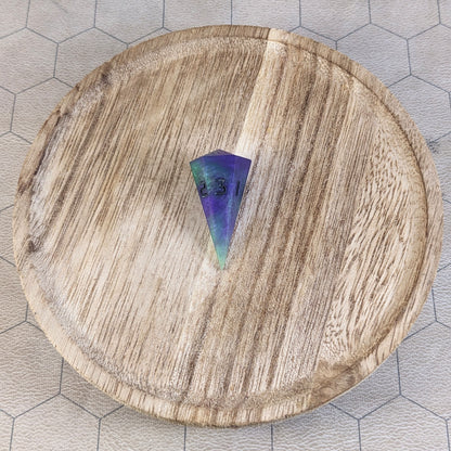 A high angle view of a dagger d3 with swirls of purple and green with black numbers that glow in the dark. It is sitting in the center of a wooden circle.