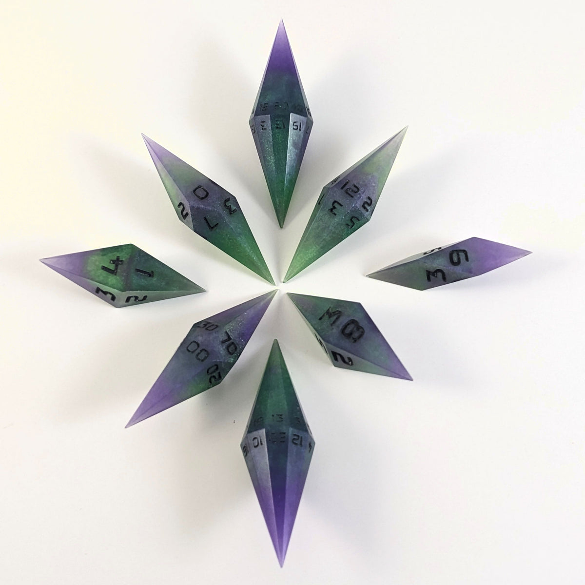 A complete set of diamond shaped resin dice featuring 2 d20s. These handmade dice are purple and green with black numbers that glow in the dark and are clustered in a circle with the bottom points angled toward a central point on a white background.