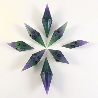 A complete set of diamond shaped resin dice featuring 2 d20s. These handmade dice are purple and green with black numbers that glow in the dark and are clustered in a circle with the bottom points angled toward a central point on a white background.