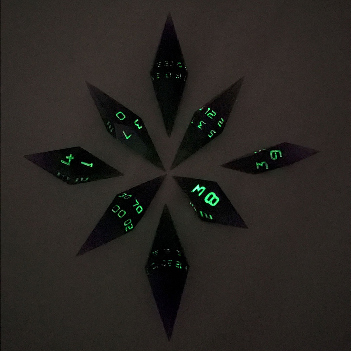 A complete set of diamond shaped resin dice featuring 2 d20s. These handmade dice are purple and green with black numbers that glow in the dark and are clustered in a circle with the bottom points angled toward a central point on a white background. This photo was taken in the dark to show the black numbers glowing green.