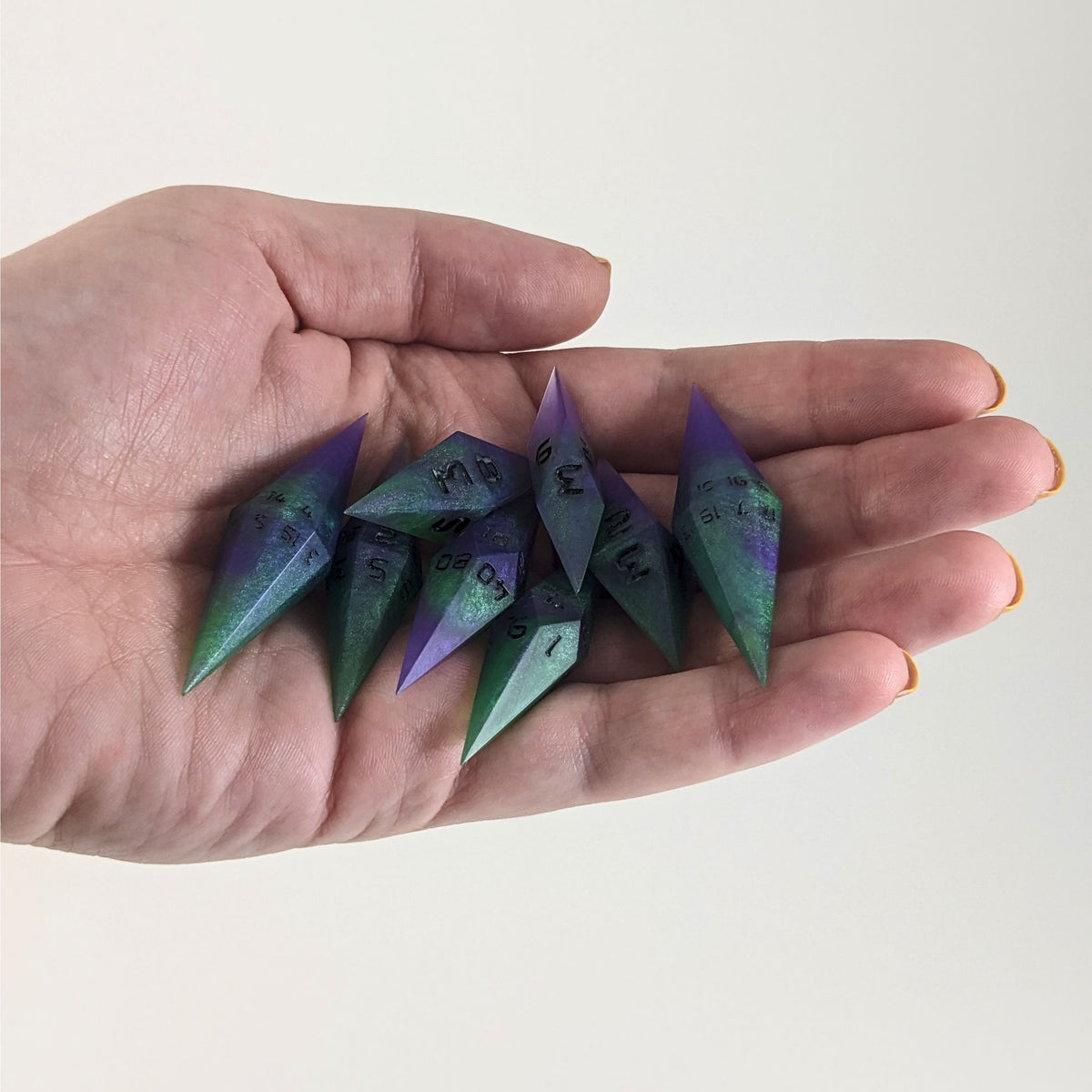 An 8 dice set of diamond style dice that are green and purple with black numbers that glow in the dark are held in a hand. The image shows that the set fits easily in a hand, making them overall smaller, though sharper, than a standard set of dnd dice.
