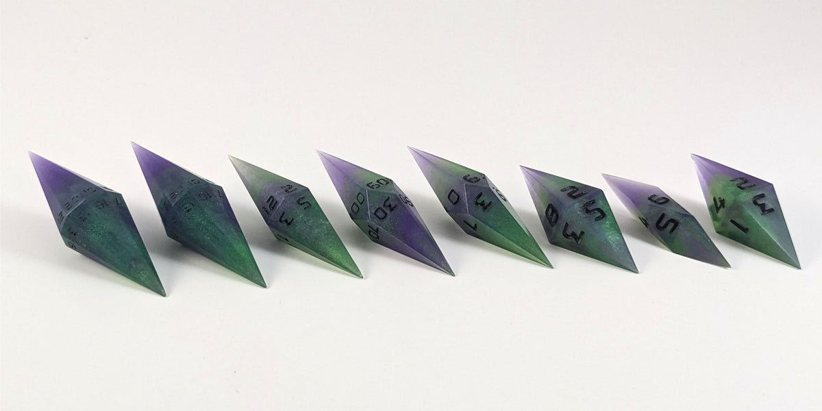 A mid angle photo of a complete set of handmade diamond DnD dice that are purple and green with black numbers that glow in the dark arranged in a straight line with bottom points angled slightly forwards and towards the right. They are lined up with the 2 d20s on the left going down to the d4 on the right side. The highest value number of each die is facing forward.