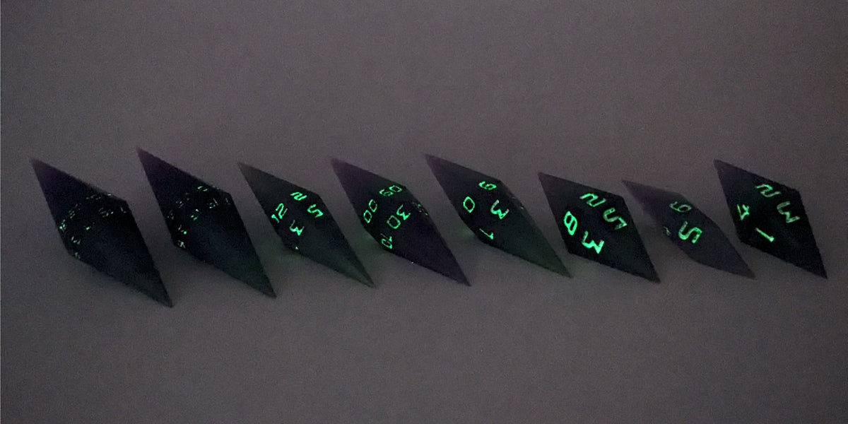 A mid angle photo of a complete set of handmade diamond DnD dice that are purple and green with black numbers that glow in the dark arranged in a straight line with bottom points angled slightly forwards and towards the right. They are lined up with the 2 d20s on the left going down to the d4 on the right side. The highest value number of each die is facing forward. The photo is taken in the dark to show the black numbers glowing green.